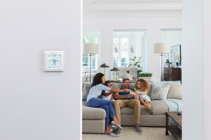 A family sits in their living room with a Honeywell Home Smart Thermostat on the wall in the foreground