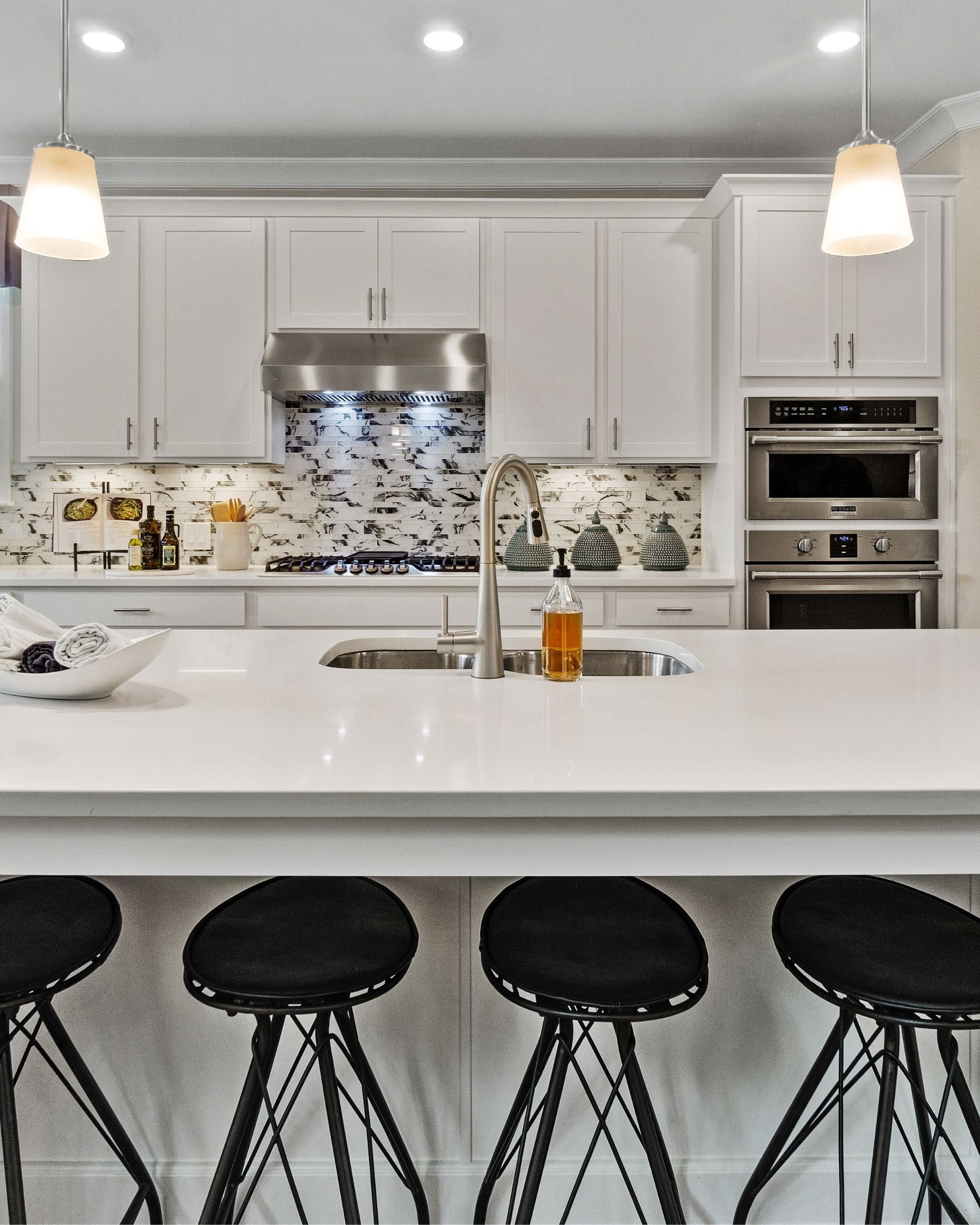 A white kitchen wtih black-speckled backsplash. A stove with hood vented to the exterior, double basin sink, and double ovens are in frame.