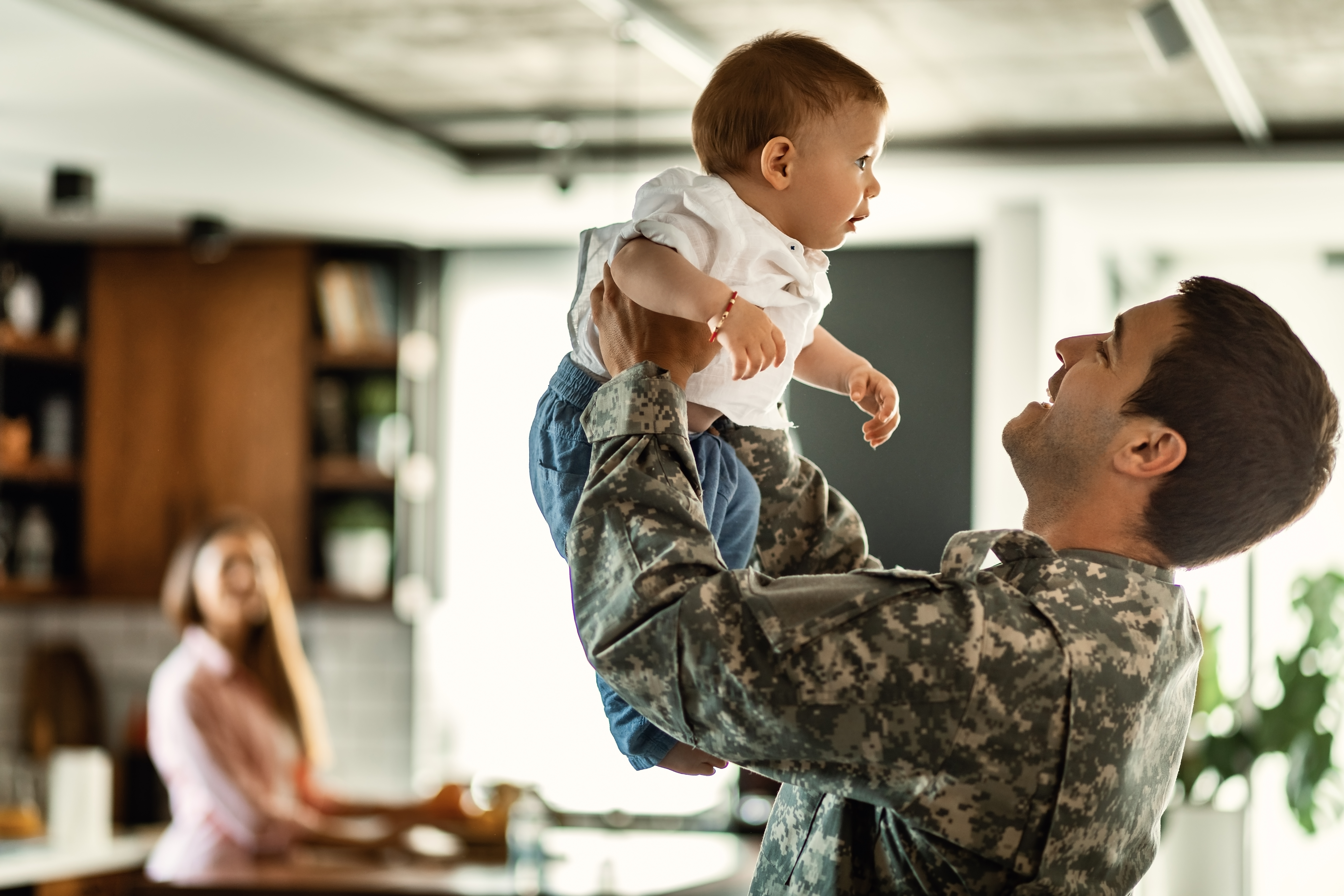 NATL_Pic_Military_Man_Holding_Up_Baby_Mom_Background_Stock_Adobe_298269578