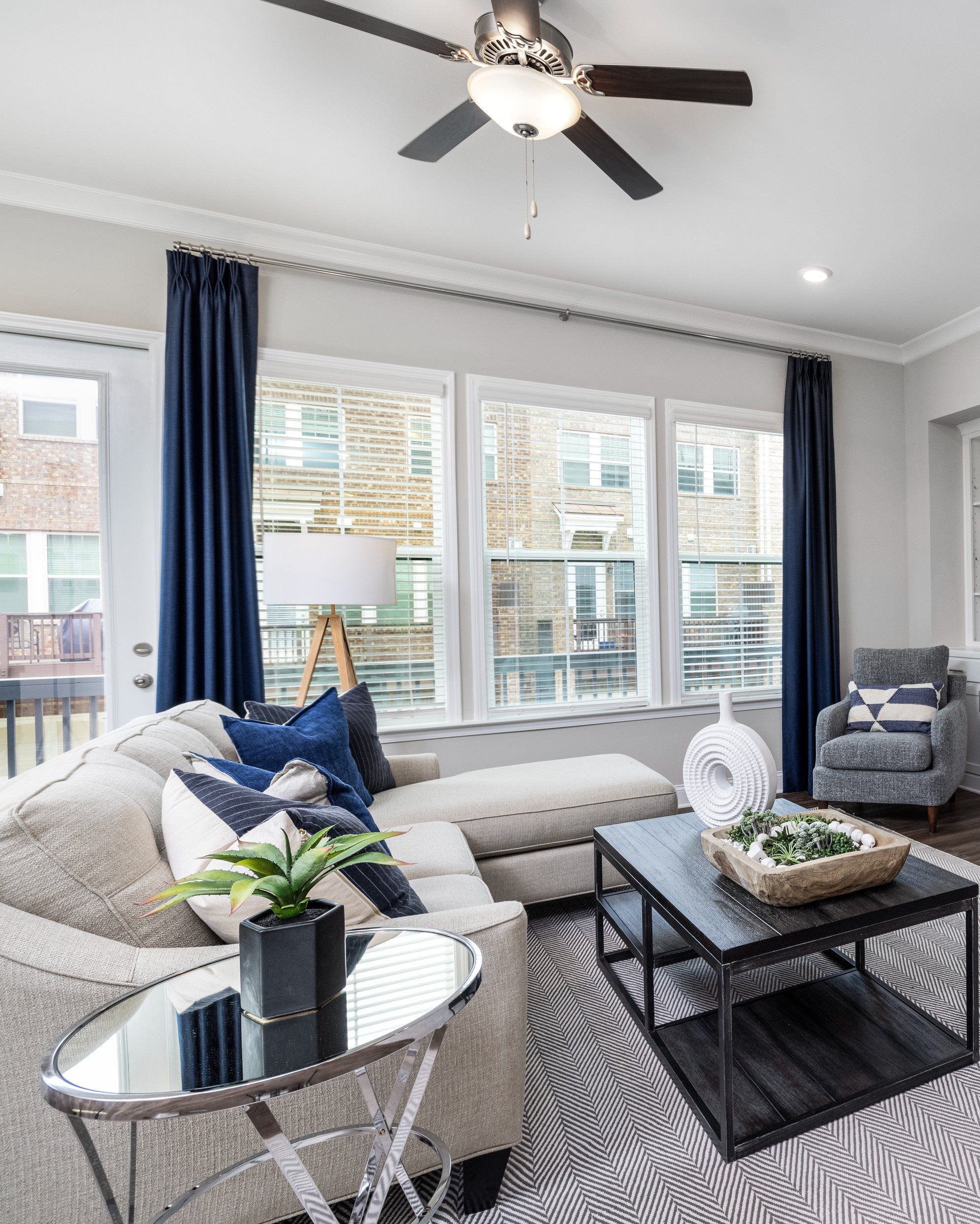 White living room with accents of blue. Windows line the living room wall, allowing in ample sunlight and offering views of the community.